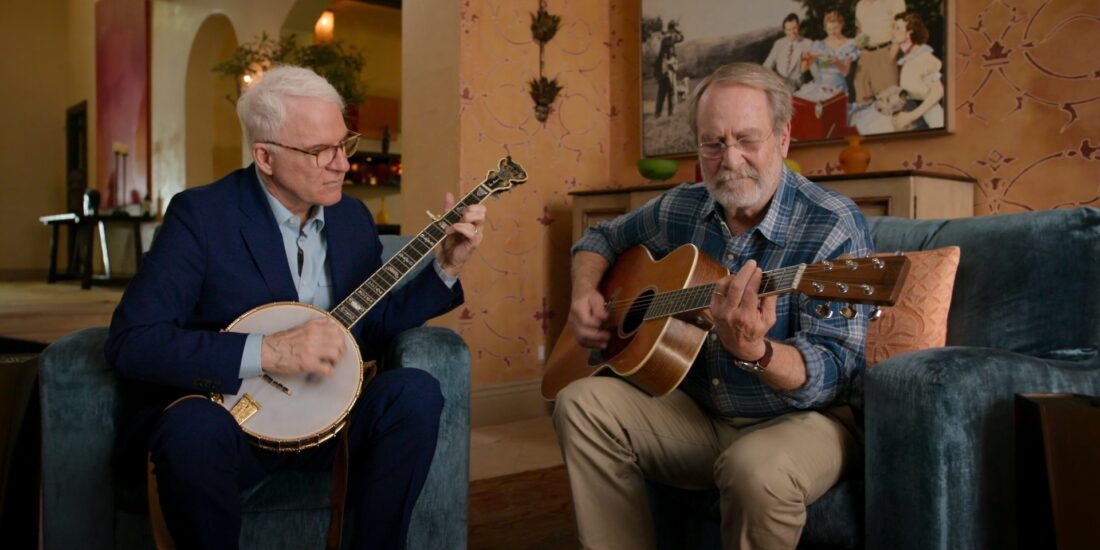 "Two older men play banjo and guitar on blue chairs in a warmly lit living room with art and a family photo on the wall behind them."